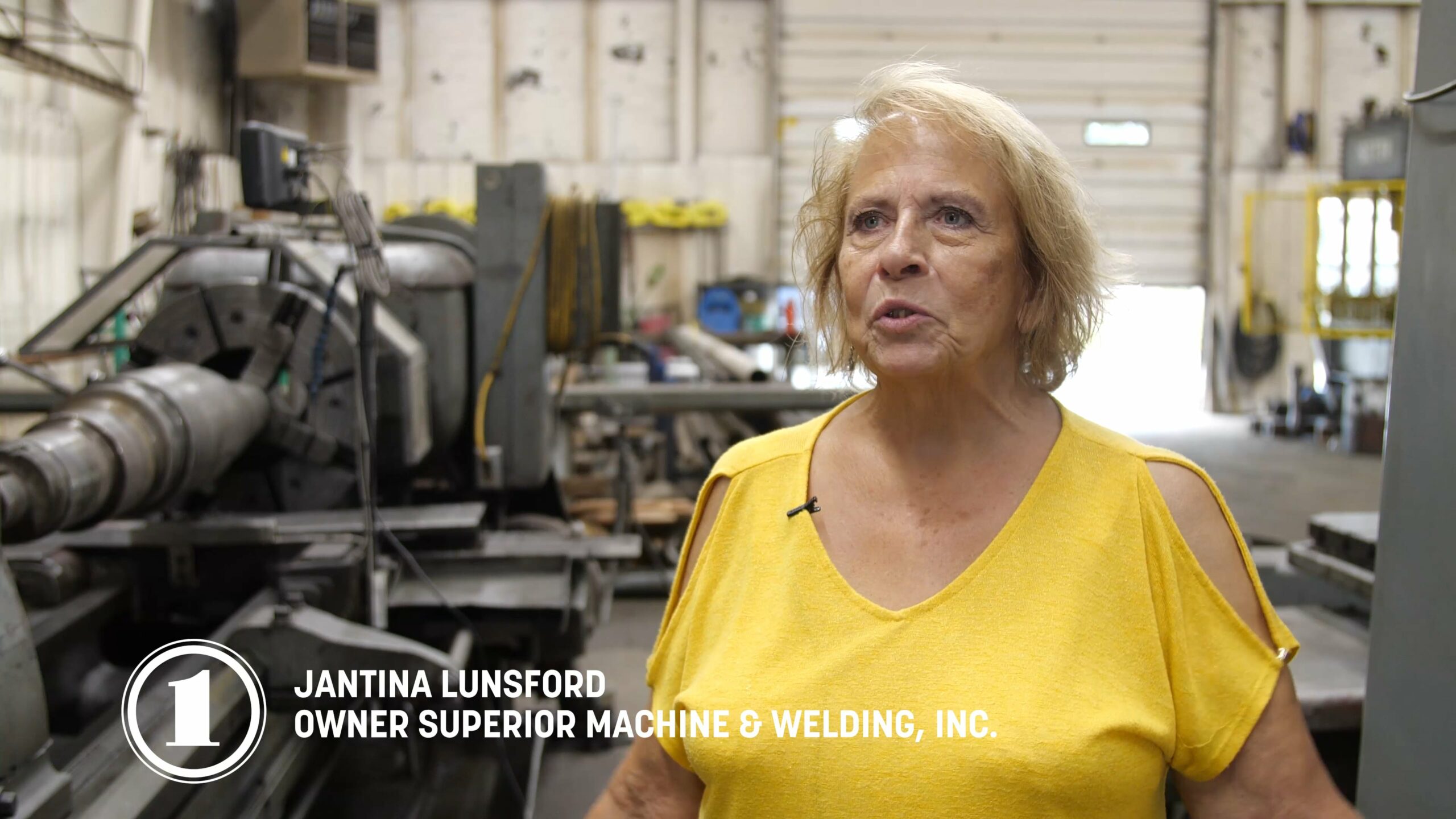 A woman wearing a yellow shirt stands confidently in front of a large machine, showcasing her professional environment.