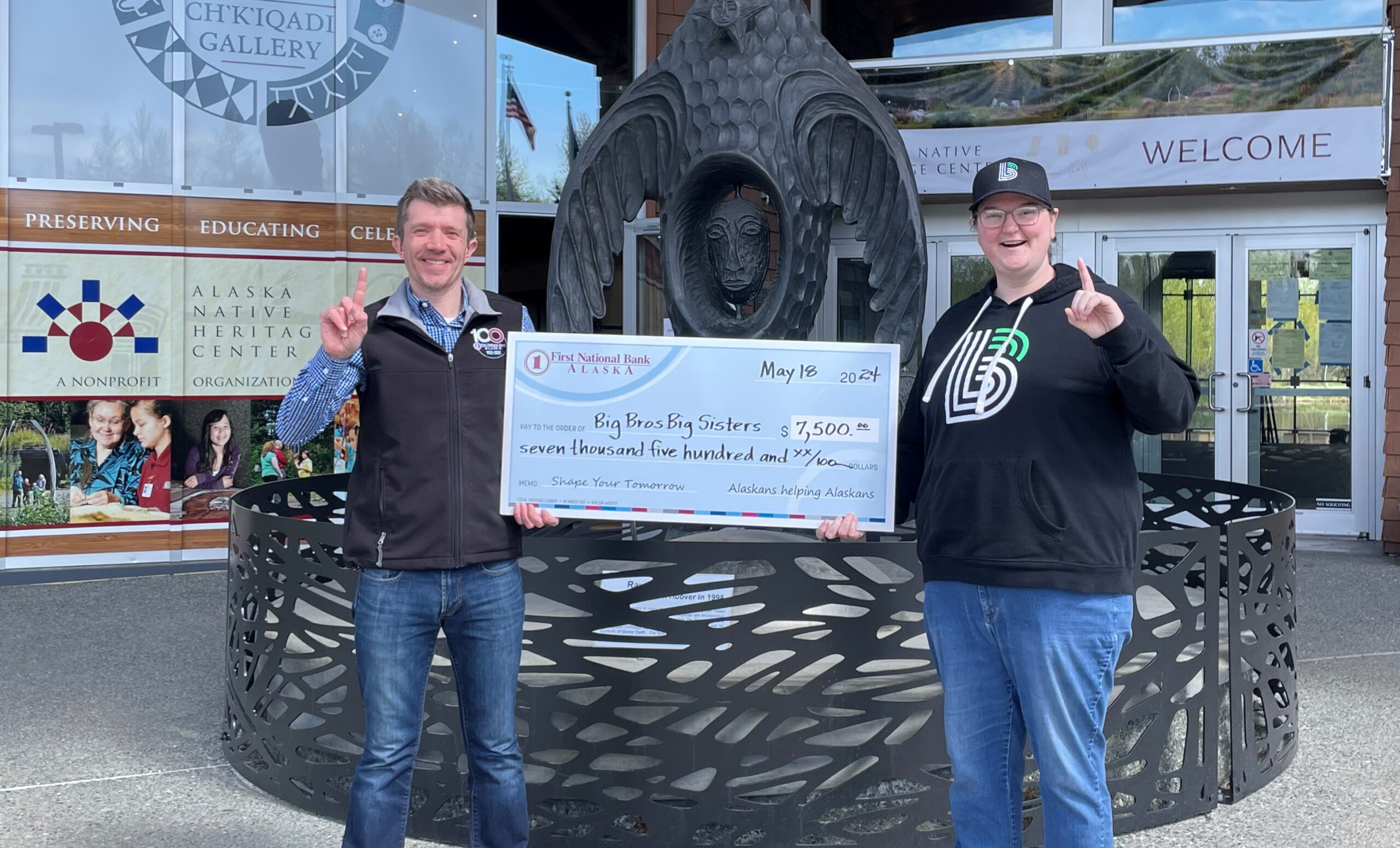 Two men proudly display a large check in front of a statue, capturing a moment of celebration and achievement.