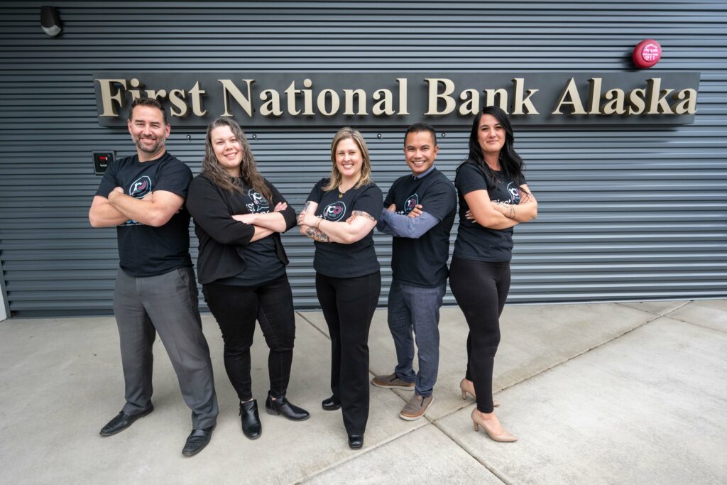 Five people stand with arms crossed and smile in front of the First National Bank Alaska building. They are wearing matching shirts and pose confidently.