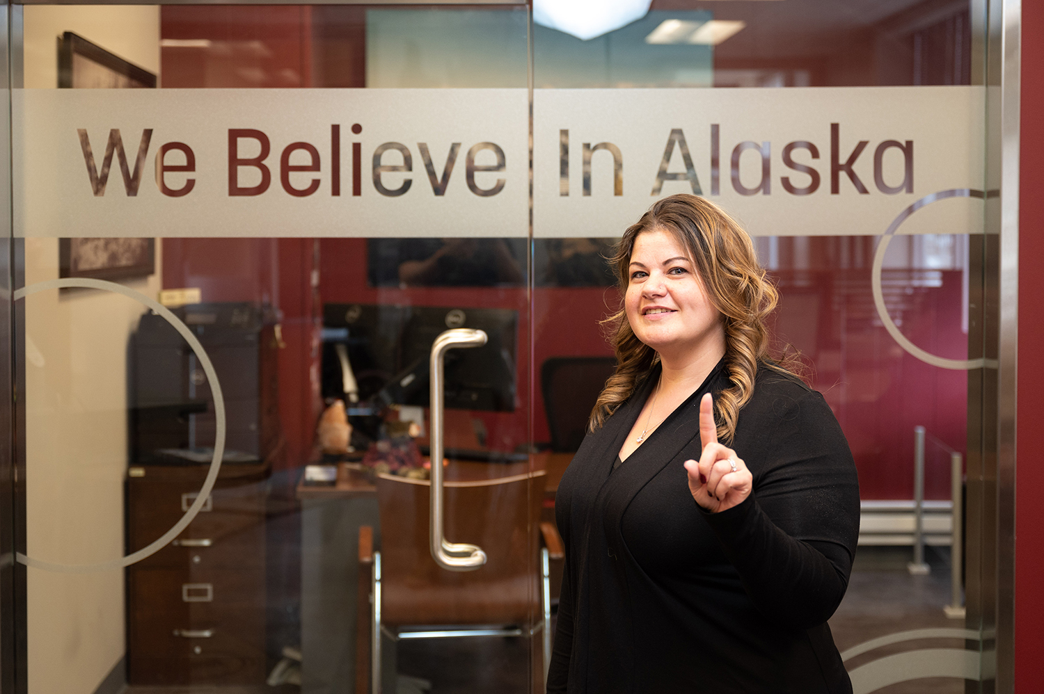 A woman poses in front of a glass door that reads "We Believe in Alaska," reflecting a message of support and unity.