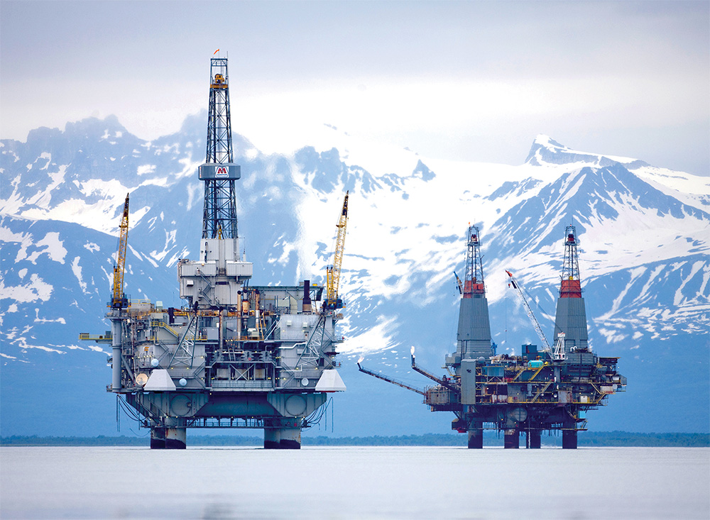 Oil rigs stand in the water, framed by majestic mountains in the background, showcasing a blend of industry and nature.